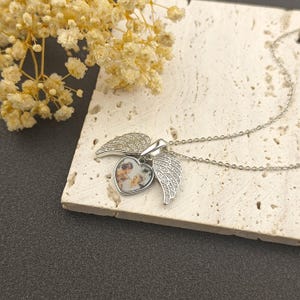 Op de afbeelding: Een zilverkleurige ketting met een hartvormige hanger met een afbeelding van twee personen. De hanger wordt geflankeerd door twee engelen vleugels, elk versierd met kleine, sprankelende stenen. De ketting ligt op een lichtgekleurd, getextureerd oppervlak.