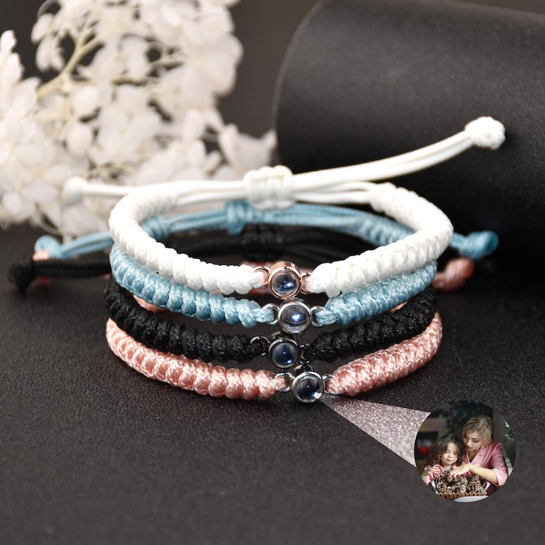 May include: Four friendship bracelets with a small, clear, round charm with a blue center. The bracelets are white, light blue, black, and pink.