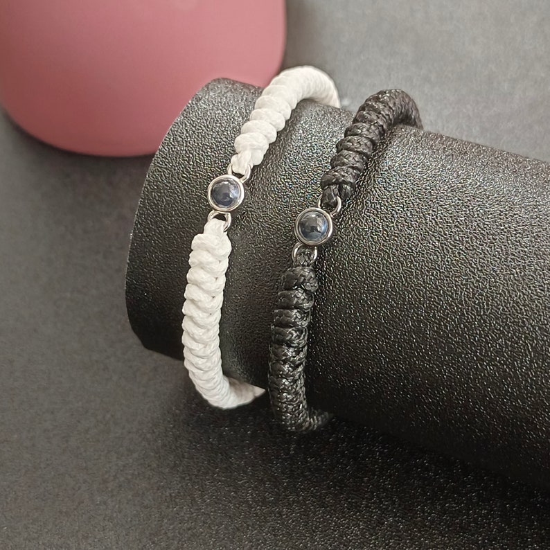 May include: Two braided bracelets with dark blue gemstone accents. One bracelet is black and the other is white.