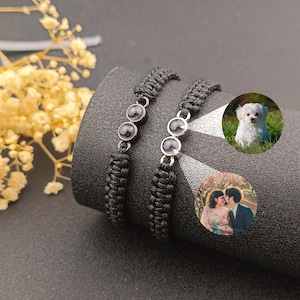 May include: Two black braided bracelets, each with three dark grey stones and silver-coloured metal accents. The bracelets are displayed with two small circular images, one of a white dog and the other of a couple.