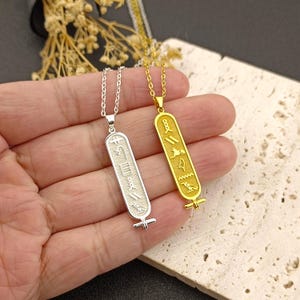 Könnte beinhalten: Zwei Anhänger-Halsketten mit rechteckigen Anhängern mit Hieroglyphen-Designs. Eine Halskette ist silberfarben, die andere goldfarben. Beide Anhänger hängen an filigranen Ketten. Die Halsketten werden vor einem neutralen Hintergrund präsentiert.