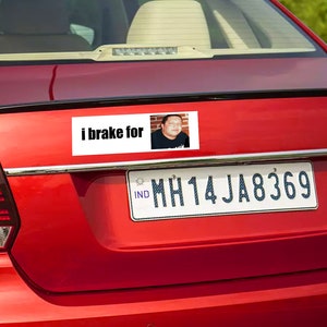 May include: A red car with a bumper sticker that says "I brake for" with a photo of a man in the sticker. The car has a license plate that says "MH14JA8369".