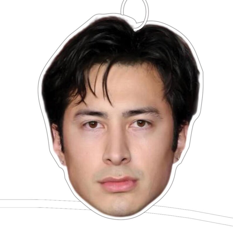 May include: A face-shaped air freshener featuring a man with dark hair and brown eyes. The air freshener has a white border and a silver loop for hanging. The man's face is the central focus of the design.
