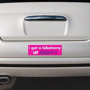 May include: A pink bumper sticker with the text "i got a lobotomy at claire's" in purple text with two pink hearts on either side of the text.