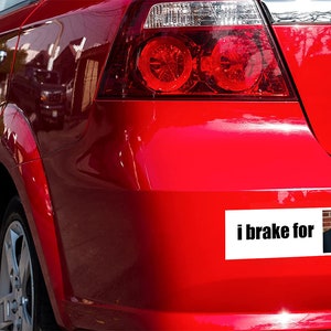 May include: A red car with a bumper sticker that says "i brake for" with a picture of a man in a black shirt.