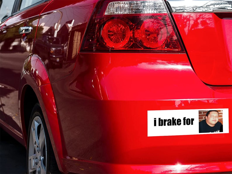 May include: A red car with a bumper sticker that says "i brake for" with a picture of a man in a black shirt.