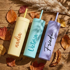 May include: Three cylindrical tumblers with matching straws in pastel yellow, mint green, and lavender. Each tumbler features a custom name in cursive script: 'nadia' in brown, 'Victoria' in teal, and 'Frieda' in black, arranged vertically.