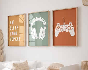 May include: Three framed wall art prints with a gaming theme. One print says "EAT SLEEP GAME REPEAT" in white text on an orange background. The other two prints feature a white headset and a white game controller on green and orange backgrounds.