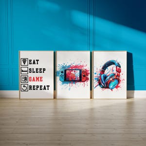 May include: Three framed art prints with a gaming theme. One print reads "EAT SLEEP GAME REPEAT". The other two prints feature a game controller and headphones, with red and blue paint splatter designs. The frames are gold.