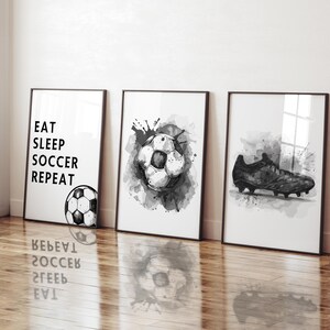 May include: Three framed black and white soccer-themed art prints. One print reads "EAT SLEEP SOCCER REPEAT" with a soccer ball illustration. The other two prints feature soccer ball and cleat illustrations with watercolor-style splatters.