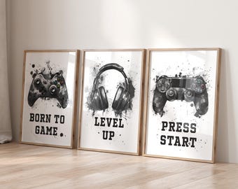 Puede incluir: Tres impresiones artísticas enmarcadas con temática de videojuegos. Cada impresión presenta un diseño de acuarela en blanco y negro de un mando de juego, auriculares y un mando, con los textos "BORN TO GAME", "LEVEL UP" y "PRESS START".