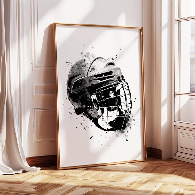 May include: Black and white art print of a hockey helmet. The artwork features a detailed illustration of the helmet with a cage, rendered in a grayscale watercolor style. The print is framed in a gold-colored frame and leans against a white wall.