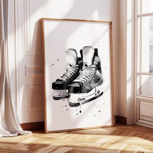 May include: Framed watercolor print of ice skates in black and white. The artwork features a pair of skates with white laces and blades, set against a white background with ink splatter details. The frame is gold.