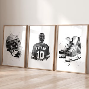 May include: Three framed black and white watercolor prints featuring hockey equipment. The prints depict a hockey helmet, a jersey with "NATHAN 10" on the back, and a pair of ice skates. The artwork has a splattered paint effect.