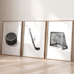 May include: Set of three framed art prints featuring watercolor illustrations of hockey equipment. Includes a black hockey puck, a hockey stick, and a hockey net. The prints are in neutral tones with wooden frames.