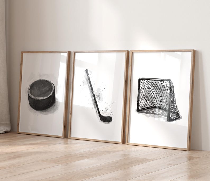 May include: Set of three framed art prints featuring watercolor illustrations of hockey equipment. Includes a black hockey puck, a hockey stick, and a hockey net. The prints are in neutral tones with wooden frames.