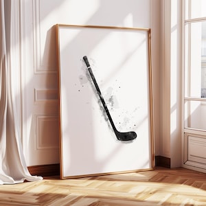 May include: A framed art print featuring a watercolor illustration of a hockey stick in black and gray. The artwork is set against a white background, with a gold frame. The print is leaning against a wall.