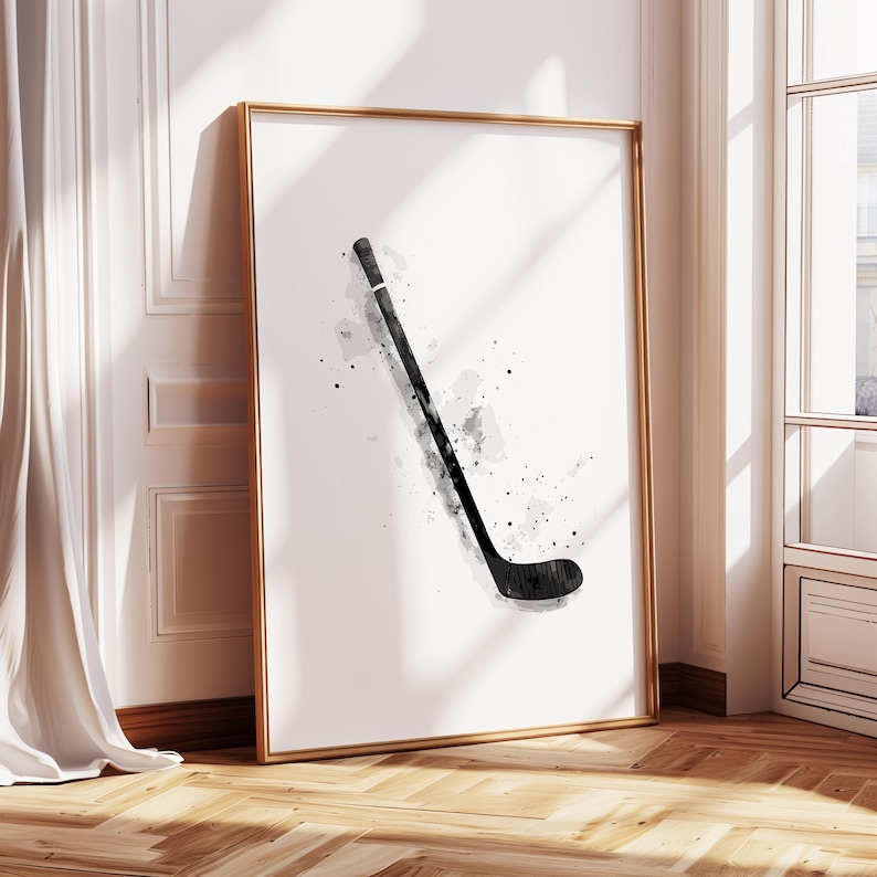 May include: A framed art print featuring a watercolor illustration of a hockey stick in black and gray. The artwork is set against a white background, with a gold frame. The print is leaning against a wall.