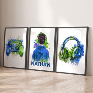 May include: Three framed watercolour art prints with gaming themes. One print shows a blue and green game controller. Another depicts a person wearing headphones, and the third shows a pair of headphones. The name "NATHAN" is printed on one of the prints.