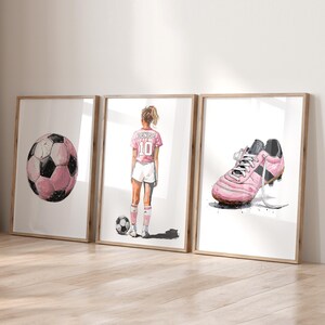 以下が含まれることがあります： サッカーをテーマにしたイラストが描かれた、額装された3つのプリント。ピンクと黒のサッカーボール、背中に「Denise」と10番のピンクのジャージを着たサッカー選手、ピンクのサッカースパイクが描かれています。白い背景にアートワークが配置されています。