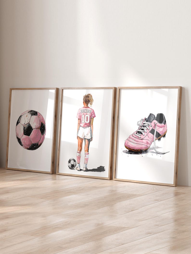 以下が含まれることがあります： サッカーをテーマにしたイラストが描かれた、額装された3つのプリント。ピンクと黒のサッカーボール、背中に「Denise」と10番のピンクのジャージを着たサッカー選手、ピンクのサッカースパイクが描かれています。白い背景にアートワークが配置されています。