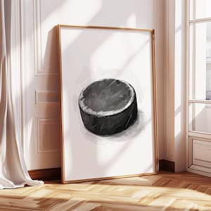 May include: A framed print featuring a watercolor illustration of a black hockey puck. The puck is depicted with shading and texture, set against a white background. The frame is gold-colored, and the artwork leans against a wall.