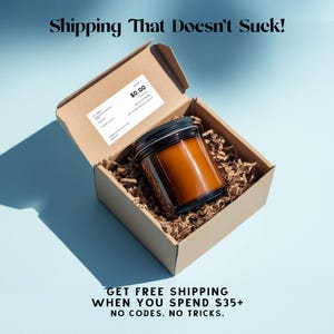 May include: A brown candle in a glass jar with a black lid sits inside an open cardboard box filled with brown packing material. The box has a shipping label with a $0.00 price. Text at the top says "Shipping That Doesn't Suck!"