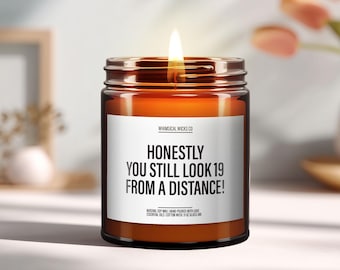 May include: A lit candle in a glass jar with a white label. The label reads "HONESTLY YOU STILL LOOK 19 FROM A DISTANCE!". The candle is amber-colored and has a cotton wick. The candle is a natural soy wax with essential oils.