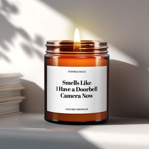 May include: A 9 oz soy candle in a brown glass jar with a white label featuring the text: THE WHIMSICAL WICKS CO. Smells Like I Have a Doorbell Camera Now 9 OZ SOY CANDLE | POURED WITH LOVE. The candle has a lit wick with a visible flame.