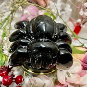 May include: A glossy black spider-shaped decorative object. The spider is resting on a small, round, gold-colored base. The background features a variety of flowers in shades of pink, white, and red.