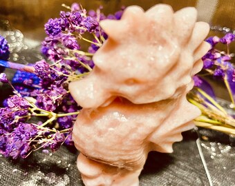 May include: A pale pink, carved seahorse figurine with a smooth, polished texture. The seahorse is positioned in front of a cluster of small, vibrant purple dried flowers, creating a contrast between the soft pink stone and the deep purple.