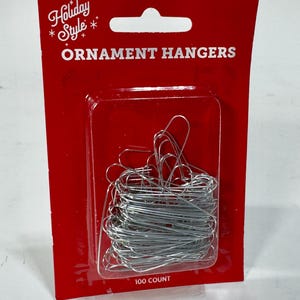 May include: Red card with a clear plastic window containing a bundle of silver ornament hangers. The card reads "Holiday Style" and "Ornament Hangers" in white text. The package contains 100 hangers.