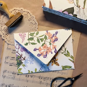May include: A white envelope with a floral pattern featuring pink, purple, and green flowers. The envelope is folded in half and has a flap on the top. The envelope is sitting on a piece of sheet music.