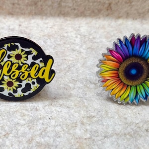 May include: Two circular pins. The left pin is black with a yellow sunflower pattern and the yellow cursive text Blessed. The right pin features a vibrant, multi-colored sunflower with a dark center and petals transitioning through the rainbow spectrum.
