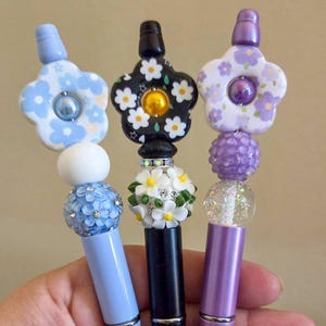May include: Three decorative pens with floral and beaded designs. The pens have light blue, black, and purple barrels. The tops feature flower-shaped accents in blue, black, and purple, with various bead embellishments. The pens are held in a hand.