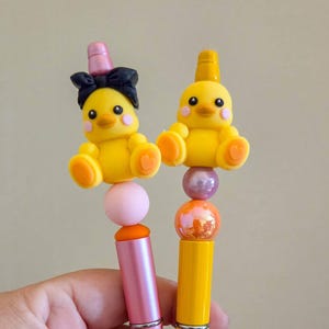 May include: Two pens featuring yellow duck toppers. The left pen is pink with a duck wearing a black bow. The right pen is yellow with a plain duck topper. Both pens include decorative beads in pink, orange, and metallic purple colors.