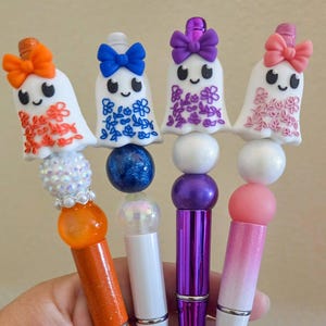 May include: Four pens featuring ghost-shaped toppers with colorful bows and floral patterns. Each pen has a unique color scheme: orange, blue, purple, and pink, accented with decorative beads in matching shades and glittery or metallic finishes.