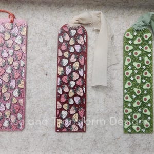 May include: Three decorative bookmarks with colorful patterns. One pink bookmark features a strawberry design and a red tassel. Another bookmark is burgundy with strawberries, and the third is green with avocado designs.