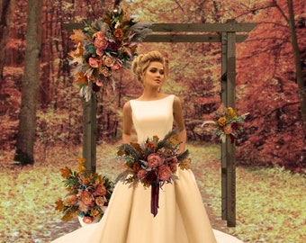 May include: A bride in a white satin wedding dress holds a bouquet of fall-colored flowers. The bouquet includes burgundy, orange, and cream flowers with greenery. The bride is standing in front of a wooden archway decorated with fall-colored flowers.