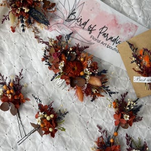 May include: Assortment of dried flower hair clips and combs in shades of orange, red, and brown. The clips are decorated with dried flowers and greenery, and the comb has a silver metal comb.