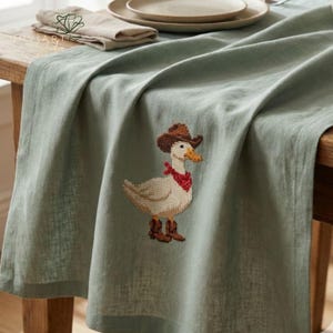 K&ouml;nnte beinhalten: Salbeigr&uuml;nes Tischtuch mit Kreuzstichmuster einer Ente mit Cowboyhut, rotem Halstuch und Stiefeln. Das Tischtuch ist &uuml;ber einem Holztisch drapiert, im Hintergrund sind eine gefaltete Serviette und Teller zu sehen.