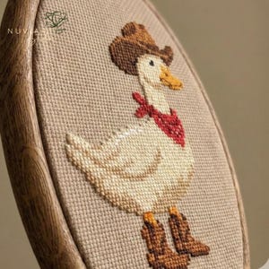 K&ouml;nnte beinhalten: Kreuzstich-Stickerei einer Ente mit Cowboyhut, rotem Halstuch und Cowboy-Stiefeln. Die Ente ist mit cremefarbenem, braunem und gelbem Garn auf einem beigefarbenen Stoff gestickt. Die Stickerei ist in einem Holzrahmen gefasst.