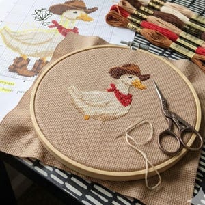 K&ouml;nnte beinhalten: Kreuzstich-Stickprojekt in einem Holzrahmen, das eine wei&szlig;e Ente mit braunem Cowboyhut und rotem Halstuch zeigt. Das Projekt befindet sich auf beigem Stoff, mit Schere, Faden und Stickgarn in verschiedenen Farben. Eine Vorlage ist sichtbar.