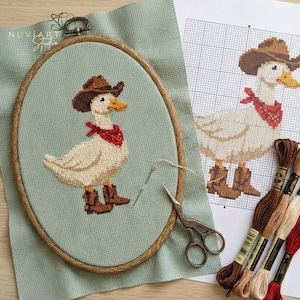 K&ouml;nnte beinhalten: Kreuzstich-Stickerei einer Ente mit Cowboyhut, rotem Halstuch und Stiefeln, in einem ovalen Holzrahmen. Das Design befindet sich auf einem hellgr&uuml;nen Stoff, mit einem Musterbogen und Stickgarn. Der Markenname "NUVIART Studio" ist sichtbar.