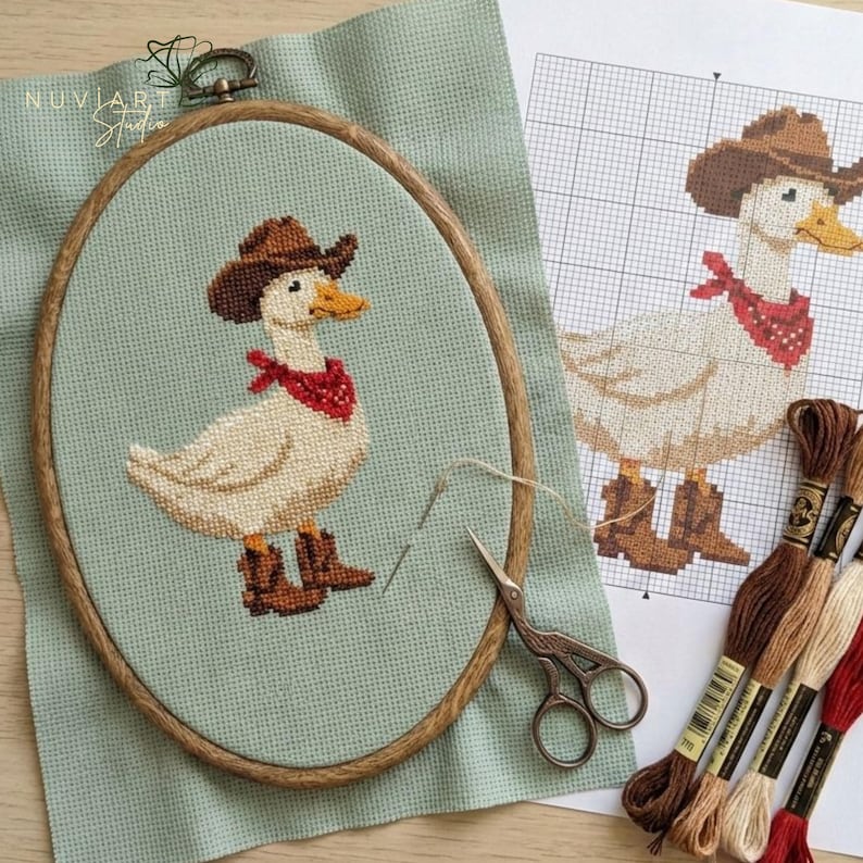 K&ouml;nnte beinhalten: Kreuzstich-Stickerei einer Ente mit Cowboyhut, rotem Halstuch und Stiefeln, in einem ovalen Holzrahmen. Das Design befindet sich auf einem hellgr&uuml;nen Stoff, mit einem Musterbogen und Stickgarn. Der Markenname "NUVIART Studio" ist sichtbar.