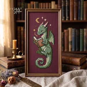 Op de afbeelding: Een ingelijst kruissteek borduurwerk met een groene draak die een open boek leest. De draak is afgebeeld met een gekrulde staart, kleine vleugels en lichtgekleurde hoorns. Boven de draak staan een gele maansikkel en kleine witte sterren.