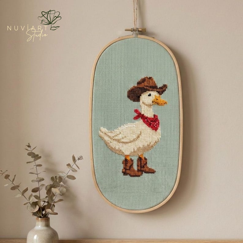 K&ouml;nnte beinhalten: Kreuzstich-Kunstwerk einer wei&szlig;en Ente mit braunem Cowboyhut, rotem Halstuch und braunen Stiefeln. Die Ente ist vor einem hellblauen Hintergrund in einem ovalen Holzrahmen platziert. Das Kunstwerk h&auml;ngt an einer Wand.
