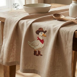 K&ouml;nnte beinhalten: Beiger Tischl&auml;ufer mit einer Stickerei einer Gans mit Cowboyhut, rotem Halstuch und Stiefeln. Der L&auml;ufer liegt auf einem Holztisch, mit einer Sch&uuml;ssel und einem L&ouml;ffel.