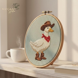 K&ouml;nnte beinhalten: Kreuzstich-Stickerei einer wei&szlig;en Ente mit braunem Cowboyhut, rotem Halstuch und braunen Stiefeln. Die Ente ist auf einem hellblauen Hintergrund zentriert und in einem Holzreifen gerahmt. Das Kunstwerk wird auf einem Holzregal ausgestellt.