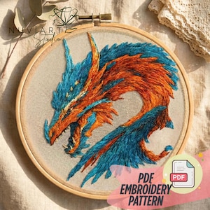Può includere: Ricamo di un drago in un telaio di legno. Il drago presenta fili blu e arancioni, con il testo "PDF EMBROIDERY PATTERN" e il logo "NUVIA ART Studio". Il ricamo è su un tessuto beige.